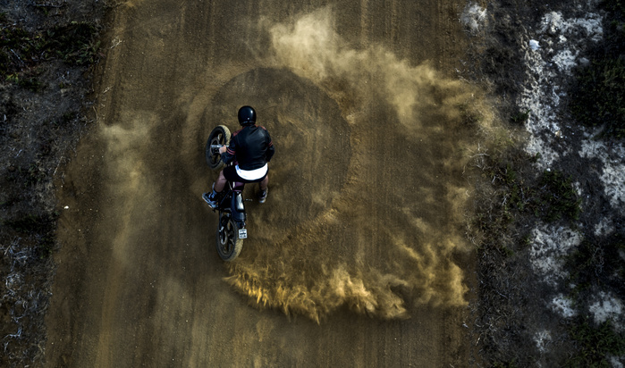 High angle view of man driving cafe racer motorcycle in circles on a dusty dirt road. High angle view of man driving cafe racer motorcycle in circles on a dusty dirt road.