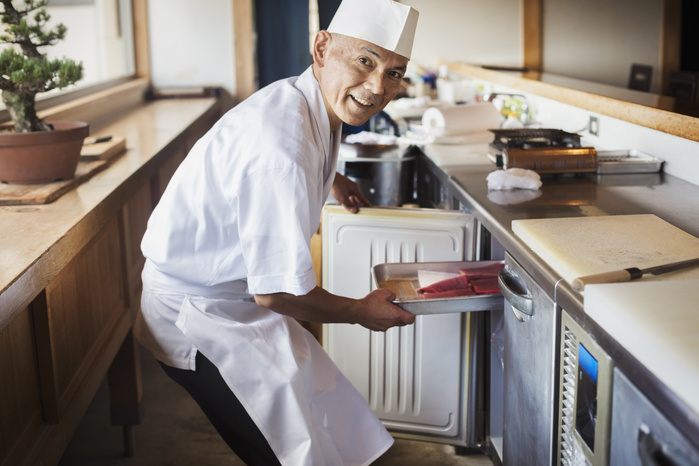 Chef working at a counter at a Japanese sushi restaurant, putting metal tray with fish in refrigerator. Chef working at a counter at a Japanese sushi restaurant, putting metal tray with fish in refrigerator.