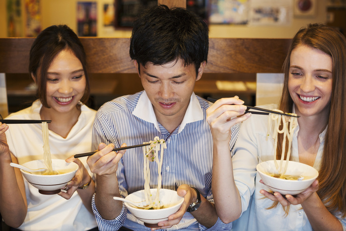 Three smiling people sitting sidy by side at a table in a restaurant, eating from bowls using chopsticks. Three smiling people sitting sidy by side at a table in a restaurant, eating from bowls using chopsticks.