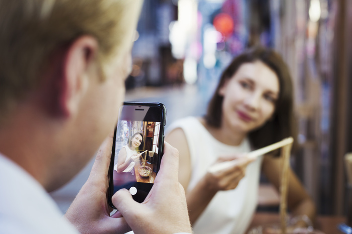 Man taking picture with smartphone of woman sitting at a table in an asian restaurant, using chopsticks, eating noodles. Man taking picture with smartphone of woman sitting at a table in an asian restaurant, using chopsticks, eating noodles.