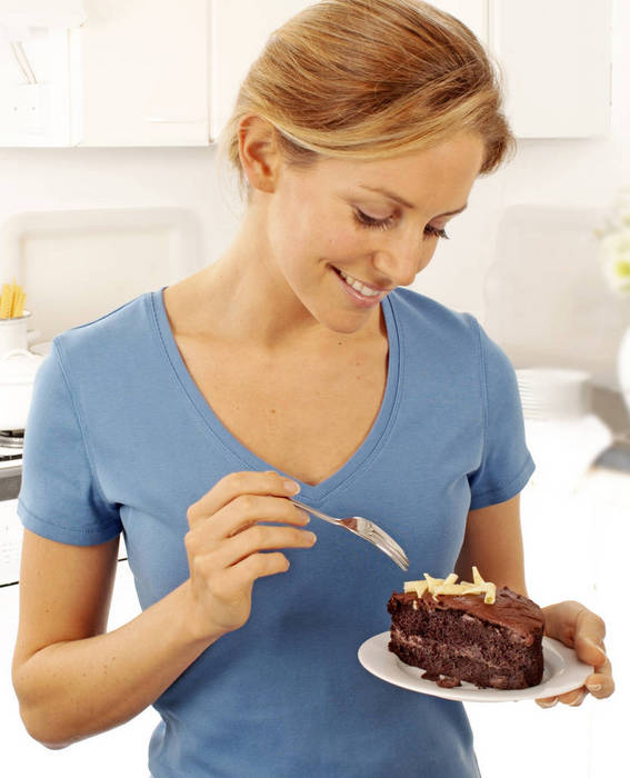 SMILING BLONDE HAIRED WOMAN IN BLUE T-SHIRT STANDING IN KITCHEN EATING SLICE OF CHOCOLATE CAKE WITH FORK