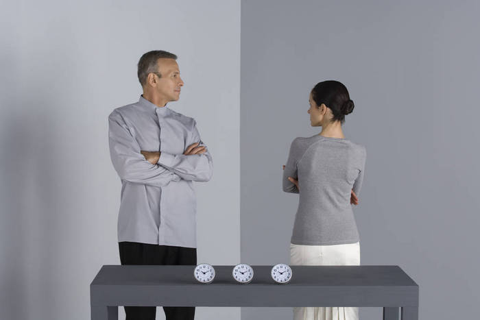 Couple standing with arms folded, looking at each other, clocks on table in front of them