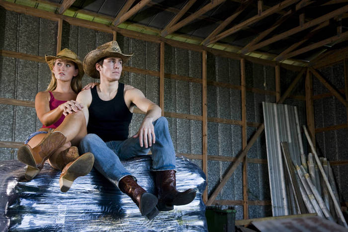 Portrait of young couple wearing cowboy hats, sitting on bags of fertilizer in a barn