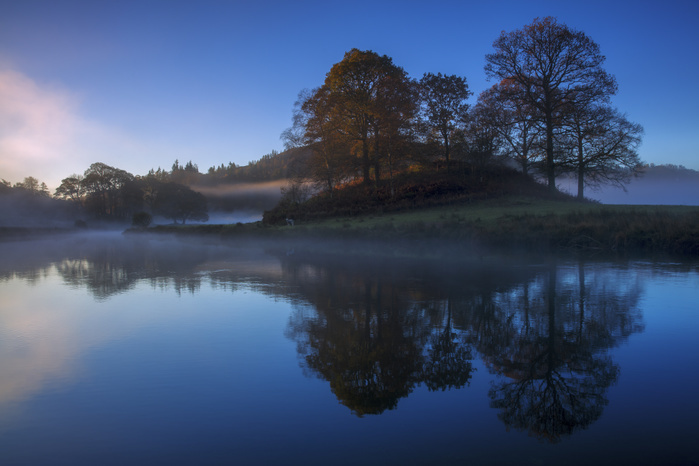 A calm, clear, autumn morning at Elterwater as the first light of a new day illuminates the trees and mist. A calm clear autumn morning at Elterwater as the first light of a new day illuminates the trees and mist, Lake District National Park, UNESCO World Heritage Site, Cumbria, England, United Kingdom, Europe, Photo by Garry Ridsdale