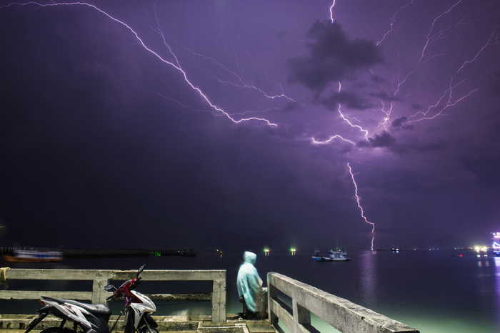 Man sitting at pier by sea against lightning in purple sky