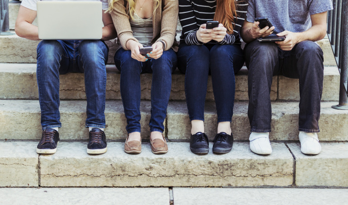 Young people looking at their cell phones. Four students sitting in a row on a step using their technology on the university campus, with a view of their feet and legs only  Edmonton, Alberta, Canada